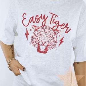 以下が含まれることがあります： 虎の顔と「Easy Tiger」という文字が赤いグラフィックで描かれた白いTシャツ。両側に稲妻が描かれている。