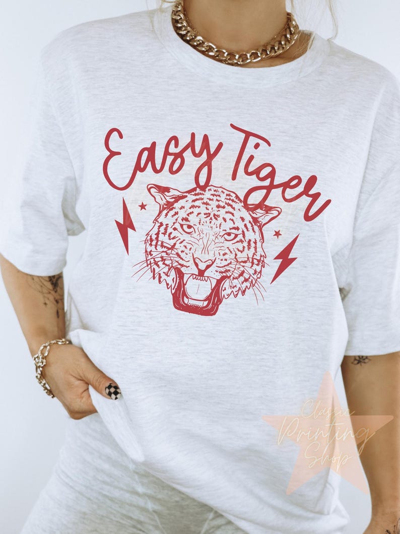以下が含まれることがあります： 虎の顔と「Easy Tiger」という文字が赤いグラフィックで描かれた白いTシャツ。両側に稲妻が描かれている。