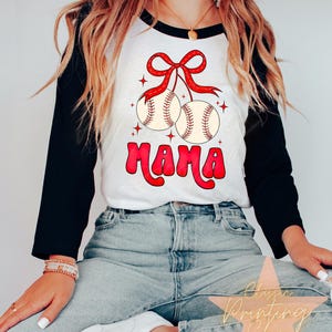 Puede incluir: Una camiseta de b&eacute;isbol blanca y negra con un lazo rojo y dos pelotas de b&eacute;isbol colgando de ella. El texto "MAMA" est&aacute; escrito en letras rojas y blancas debajo de las pelotas de b&eacute;isbol.
