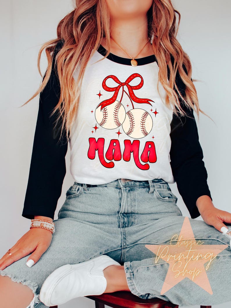Puede incluir: Una camiseta de b&eacute;isbol blanca y negra con un lazo rojo y dos pelotas de b&eacute;isbol colgando de ella. El texto "MAMA" est&aacute; escrito en letras rojas y blancas debajo de las pelotas de b&eacute;isbol.