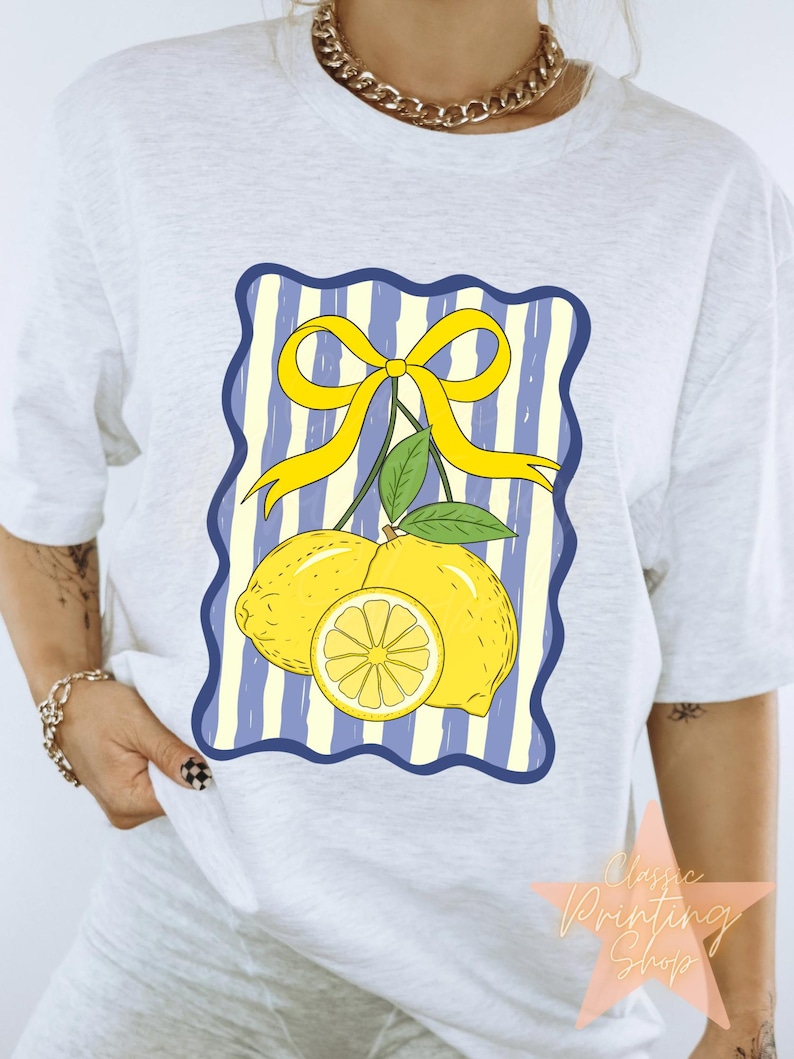 May include: A light gray t-shirt featuring a graphic of two lemons, a lemon slice, and a yellow bow on a blue and white striped background. The text "Classic Printing Shop" is visible in the lower right corner.