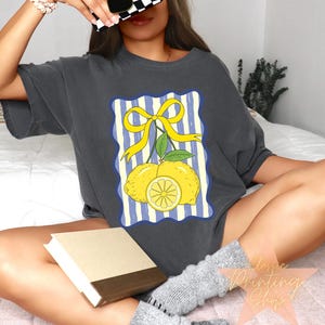 May include: Dark gray t-shirt featuring a lemon graphic. The design includes two yellow lemons, a lemon slice, and a yellow bow on a blue and white striped background. The shirt is a casual style.