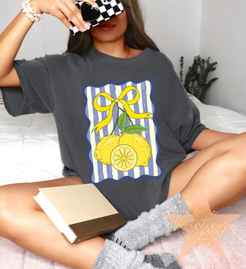 May include: Dark gray t-shirt featuring a lemon graphic. The design includes two yellow lemons, a lemon slice, and a yellow bow on a blue and white striped background. The shirt is a casual style.
