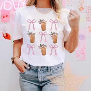 May include: White t-shirt with a graphic of multiple iced coffee cups with pink bows. The coffee cups have a brown and white design.