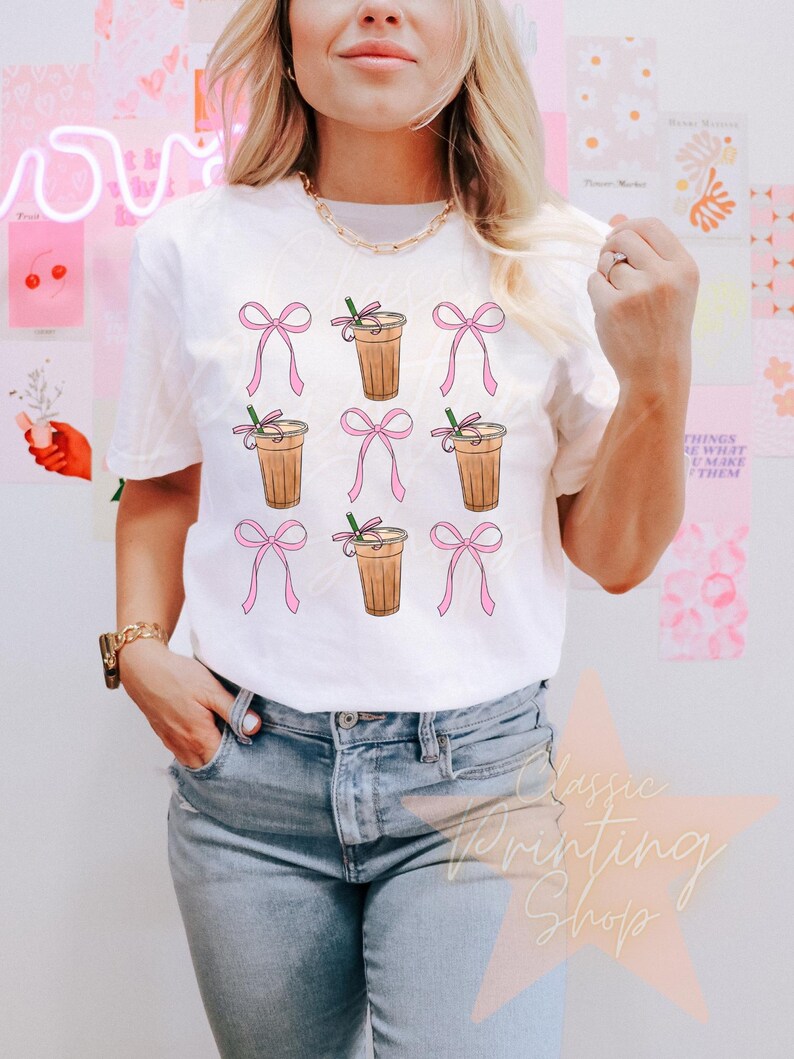 May include: White t-shirt with a graphic of multiple iced coffee cups with pink bows. The coffee cups have a brown and white design.