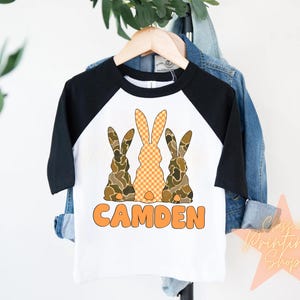 May include: A white and black baseball tee shirt with a design of three brown and orange bunnies. The text "CAMDEN" is printed in orange below the bunnies.