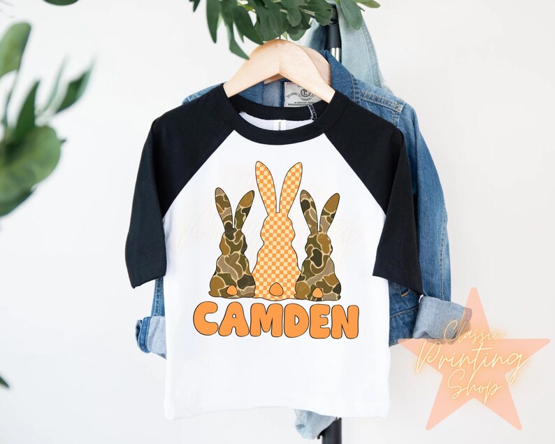 May include: A white and black baseball tee shirt with a design of three brown and orange bunnies. The text "CAMDEN" is printed in orange below the bunnies.