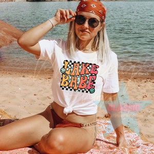 Puede incluir: Camiseta blanca con un gr&aacute;fico colorido que dice "Lake Babe" con caritas sonrientes. La camiseta est&aacute; anudada a la cintura y se lleva con una bandana roja y gafas de sol.