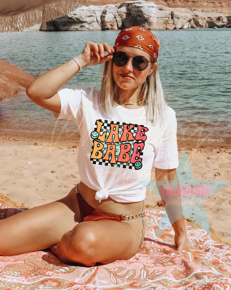 Puede incluir: Camiseta blanca con un gr&aacute;fico colorido que dice "Lake Babe" con caritas sonrientes. La camiseta est&aacute; anudada a la cintura y se lleva con una bandana roja y gafas de sol.