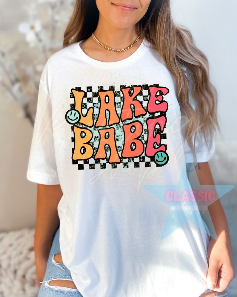 Puede incluir: Camiseta blanca con un gr&aacute;fico colorido que dice "LAKE BABE" en una fuente retro con un fondo a cuadros y caritas sonrientes.