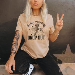 May include: Tan t-shirt with the text "Gentle Parenting Drop Out" in black and white. The graphic includes a skeleton, lightning bolt, and stars. The person is wearing black jeans and white sneakers.