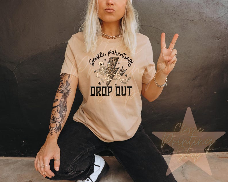 May include: Tan t-shirt with the text "Gentle Parenting Drop Out" in black and white. The graphic includes a skeleton, lightning bolt, and stars. The person is wearing black jeans and white sneakers.