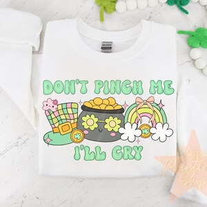 May include: White sweatshirt with a green, yellow, and pink cartoon design. The design features a pot of gold with a face, a rainbow, and a leprechaun hat. The text "Don't Pinch Me I'll Cry" is written in green letters.