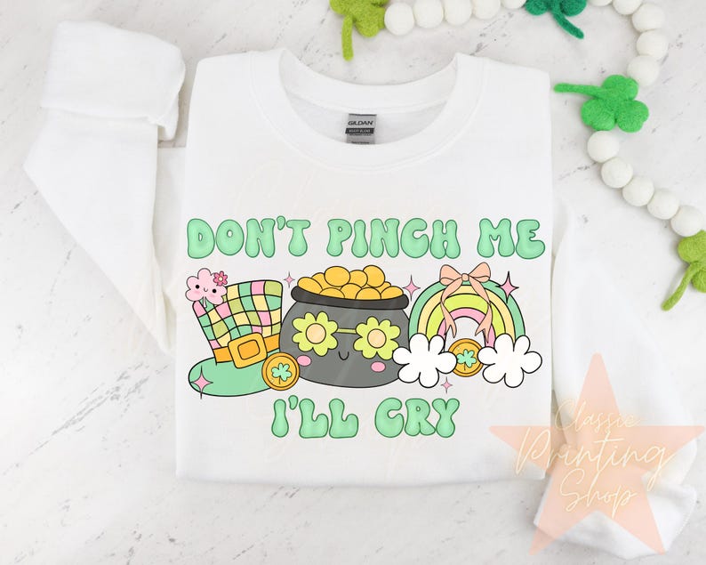May include: White sweatshirt with a green, yellow, and pink cartoon design. The design features a pot of gold with a face, a rainbow, and a leprechaun hat. The text "Don't Pinch Me I'll Cry" is written in green letters.