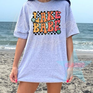 Puede incluir: Una camiseta gris con un gr&aacute;fico de estilo retro que dice "LAKE BABE" en letras de colores con caritas sonrientes a cada lado. El gr&aacute;fico est&aacute; sobre un fondo a cuadros blanco y negro.