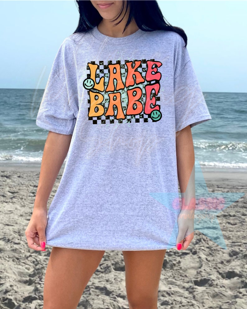 Puede incluir: Una camiseta gris con un gr&aacute;fico de estilo retro que dice "LAKE BABE" en letras de colores con caritas sonrientes a cada lado. El gr&aacute;fico est&aacute; sobre un fondo a cuadros blanco y negro.