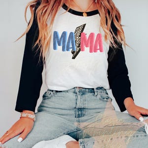 May include: A white and black baseball tee shirt with a blue and pink graphic that says "MAMA" with a leopard print lightning bolt in the middle.