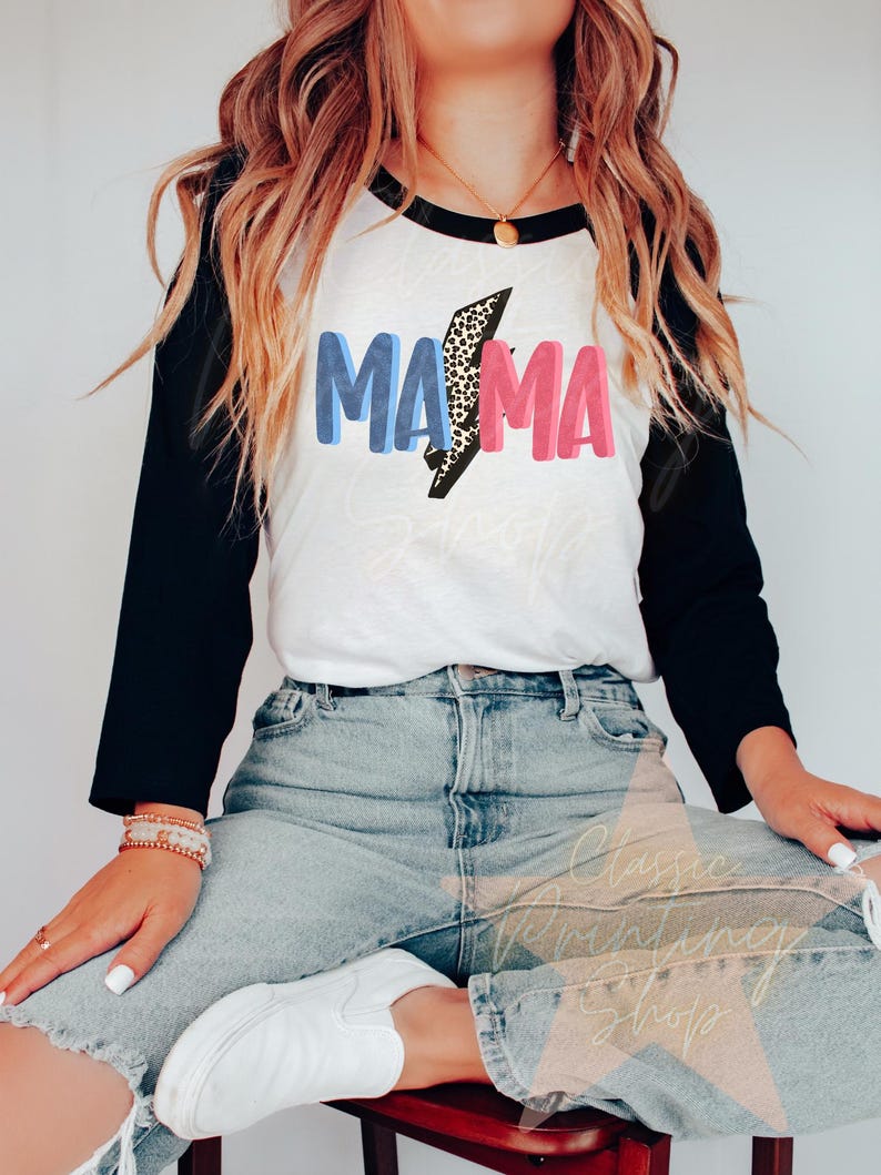 May include: A white and black baseball tee shirt with a blue and pink graphic that says "MAMA" with a leopard print lightning bolt in the middle.