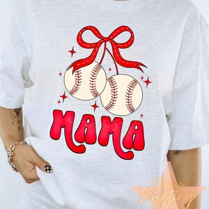 Puede incluir: Camiseta blanca con un lazo rojo y dos pelotas de b&eacute;isbol colgando de &eacute;l. La palabra "MAMA" est&aacute; escrita en rojo debajo de las pelotas de b&eacute;isbol.