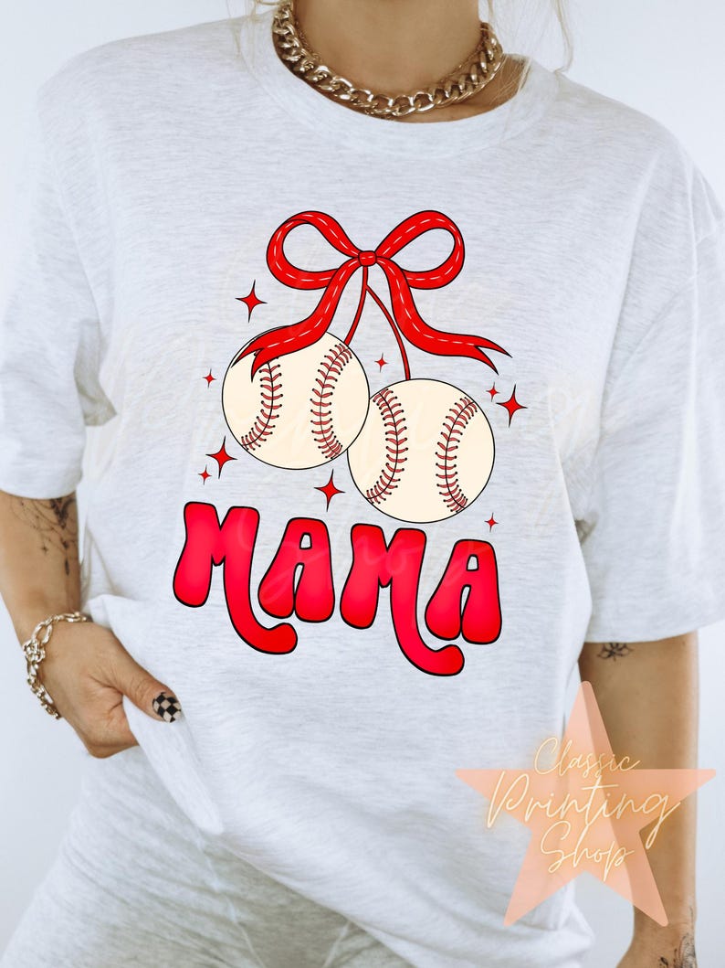 Puede incluir: Camiseta blanca con un lazo rojo y dos pelotas de b&eacute;isbol colgando de &eacute;l. La palabra "MAMA" est&aacute; escrita en rojo debajo de las pelotas de b&eacute;isbol.