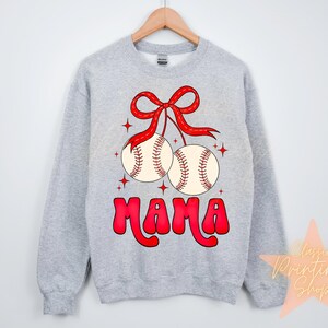 Puede incluir: Sudadera gris con un lazo rojo y dos pelotas de b&eacute;isbol colgando de ella. Las pelotas de b&eacute;isbol son blancas con costuras rojas. El texto "MAMA" est&aacute; escrito en rojo debajo de las pelotas de b&eacute;isbol.
