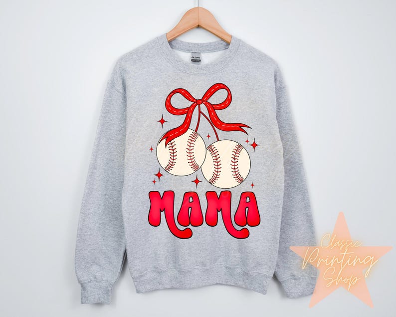 Puede incluir: Sudadera gris con un lazo rojo y dos pelotas de b&eacute;isbol colgando de ella. Las pelotas de b&eacute;isbol son blancas con costuras rojas. El texto "MAMA" est&aacute; escrito en rojo debajo de las pelotas de b&eacute;isbol.