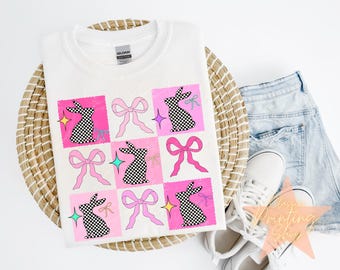 May include: A white t-shirt with a checkered bunny rabbit design. The design is made up of nine squares, each with a different color and pattern. The squares are arranged in a 3x3 grid. The bunny rabbits are all wearing pink bows.