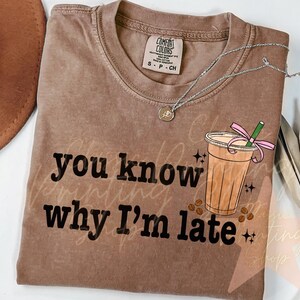 Puede incluir: Una camiseta marr&oacute;n con el texto "you know why I'm late" impreso en negro. Una ilustraci&oacute;n de dibujos animados de una bebida de caf&eacute; helado con una cinta rosa y una pajita est&aacute; por encima del texto.