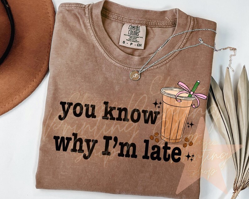 Puede incluir: Una camiseta marr&oacute;n con el texto "you know why I'm late" impreso en negro. Una ilustraci&oacute;n de dibujos animados de una bebida de caf&eacute; helado con una cinta rosa y una pajita est&aacute; por encima del texto.