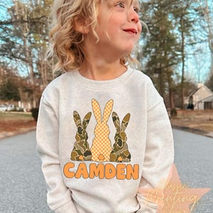 May include: A white sweatshirt with a graphic of three bunnies. The bunnies are wearing camouflage and checkered patterns. The text "CAMDEN" is printed in orange below the bunnies.
