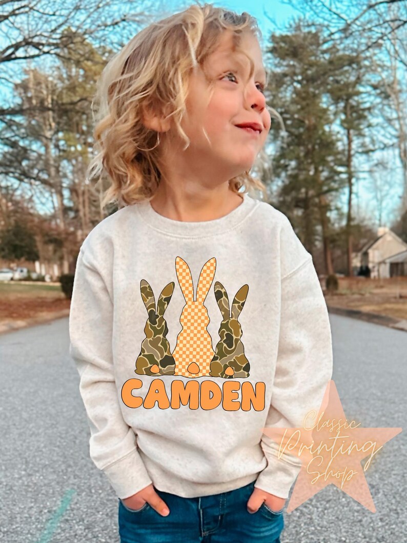 May include: A white sweatshirt with a graphic of three bunnies. The bunnies are wearing camouflage and checkered patterns. The text "CAMDEN" is printed in orange below the bunnies.