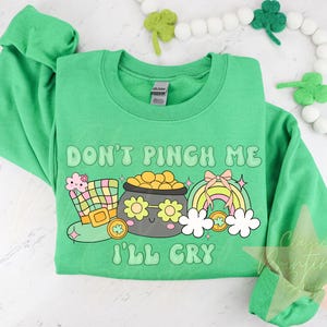 May include: Green sweatshirt with the text "Don't Pinch Me I'll Cry" in a rainbow font. The design includes a cartoon pot of gold, a leprechaun hat, and a rainbow.