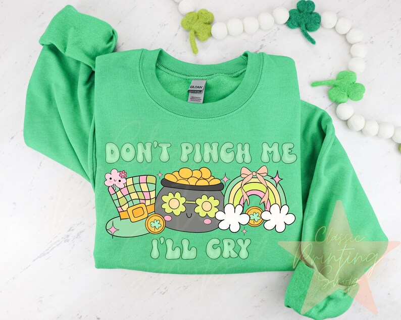 May include: Green sweatshirt with the text "Don't Pinch Me I'll Cry" in a rainbow font. The design includes a cartoon pot of gold, a leprechaun hat, and a rainbow.