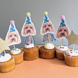 May include: Cupcakes with white icing and sprinkles, decorated with dog-themed party hats and gold stars. The cupcakes are arranged on a peach-coloured, round surface, perfect for a celebration or party.