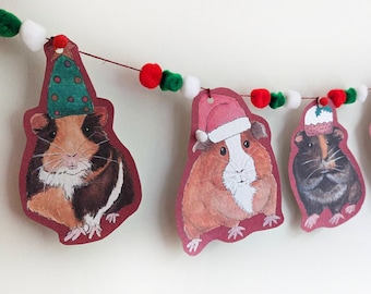 Peut inclure: Une guirlande de Noël avec des pompons rouges, verts et blancs et des cochons d'Inde festifs portant des bonnets de Père Noël et d'autres couvre-chefs de fête.