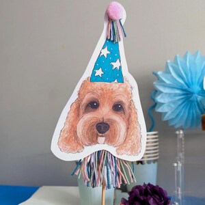 May include: A cake topper with a dog illustration wearing a blue party hat with white stars. The dog has brown fur and a sweet expression. The topper is on a wooden stick, placed on a frosted cake with red sprinkles.