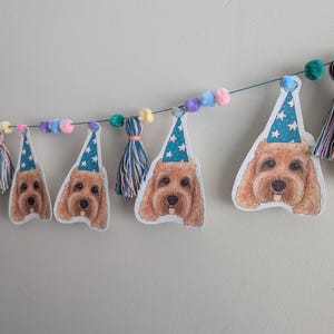 Könnte beinhalten: Ein festliches Banner mit Illustrationen von Hundegesichtern, die blaue Partyhüte mit weißen Sternen tragen. Das Banner ist mit bunten Pompons und Quasten versehen und schafft eine fröhliche und festliche Atmosphäre. Ideal für eine Hundegeburtstagsparty.