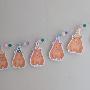 May include: A party banner with illustrations of orange capybaras wearing party hats. The banner is strung with a white string and colourful pom-poms. The capybaras are printed on white paper.