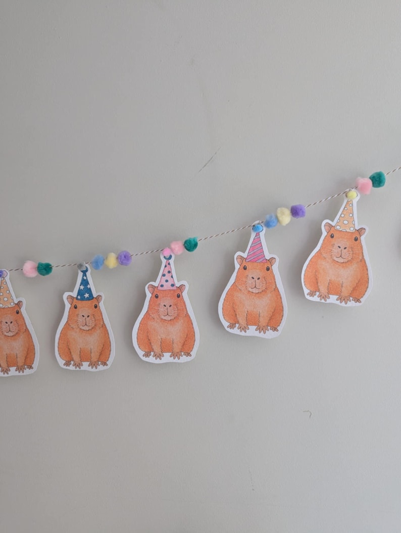 May include: A party banner with illustrations of orange capybaras wearing party hats. The banner is strung with a white string and colourful pom-poms. The capybaras are printed on white paper.