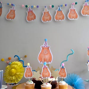 May include: A party scene featuring a garland of capybara cutouts in party hats. Cupcakes with capybara toppers are on a gold cake stand. Decorative elements include a yellow paper flower and blue accents.