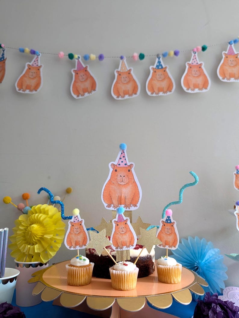 May include: A party scene featuring a garland of capybara cutouts in party hats. Cupcakes with capybara toppers are on a gold cake stand. Decorative elements include a yellow paper flower and blue accents.