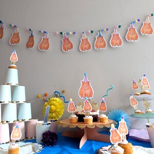 May include: A party scene with capybara decorations. A garland of capybara cutouts and pom-poms hangs above cupcakes and a tiered dessert stand. The cupcakes are topped with capybara toppers, creating a whimsical birthday celebration.