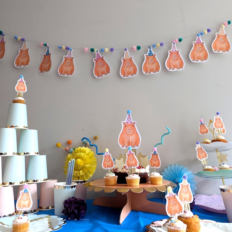 May include: A party scene with capybara decorations. A garland of capybara cutouts and pom-poms hangs above cupcakes and a tiered dessert stand. The cupcakes are topped with capybara toppers, creating a whimsical birthday celebration.