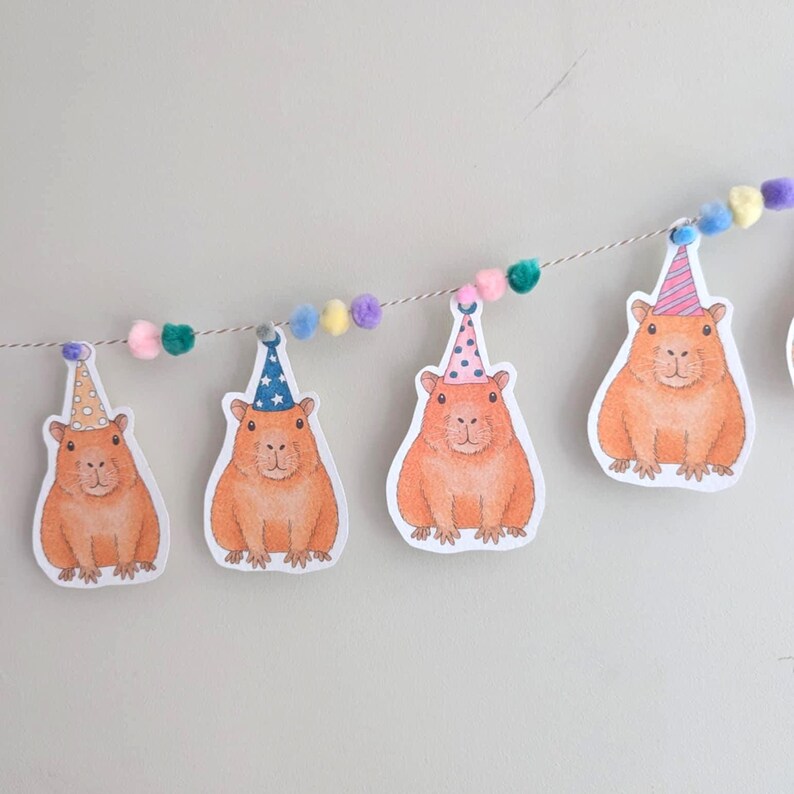 May include: A decorative garland featuring orange capybara illustrations wearing party hats. The garland is strung with a thin cord and decorated with colourful pom-poms in shades of pink, green, yellow, and purple.