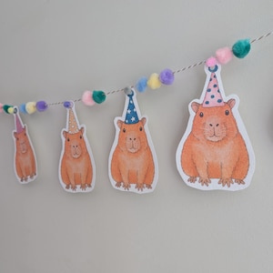 May include: A decorative garland with illustrations of orange capybaras wearing party hats. The capybaras are strung on a thin white string, with colourful pom-poms between them. The background is a plain, light grey wall.