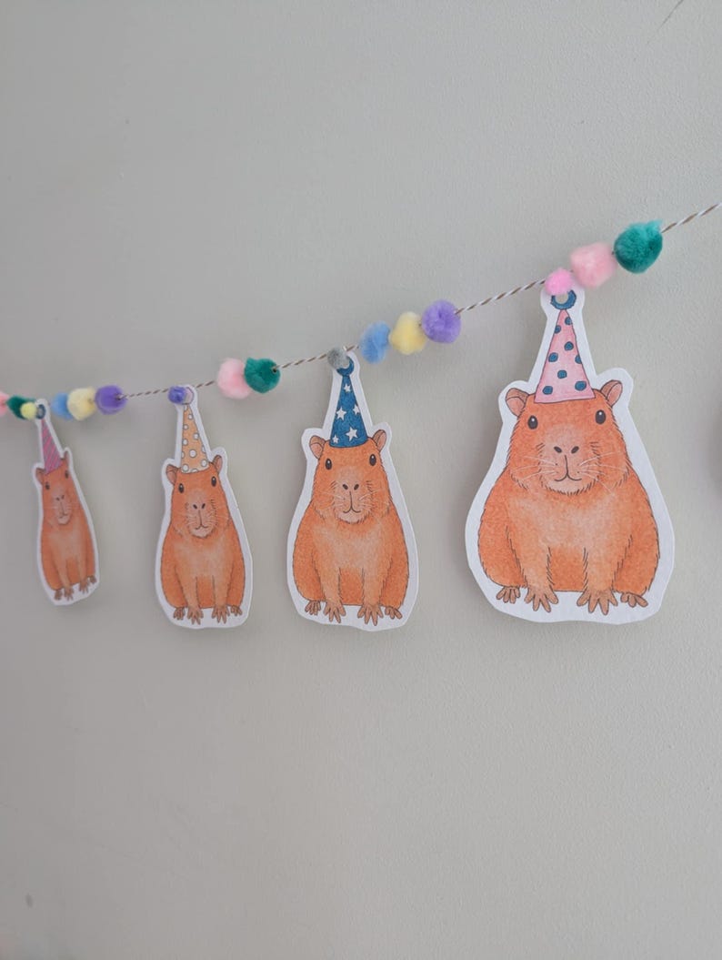 May include: A decorative garland with illustrations of orange capybaras wearing party hats. The capybaras are strung on a thin white string, with colourful pom-poms between them. The background is a plain, light grey wall.