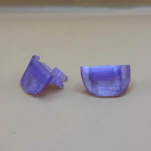 May include: Two-piece, translucent purple plastic object. One piece is a rounded shape, the other has a hinged design with a locking mechanism. The objects are on a light tan surface. The pieces appear to be 3D printed.