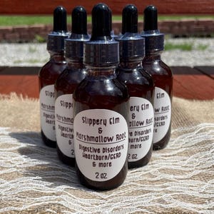 May include: Six brown glass bottles with black droppers are arranged on a beige surface. Each bottle has a white label with black text that reads "Slippery Elm & Marshmallow Root" and mentions digestive disorders and heartburn. The bottles are 2 oz each.
