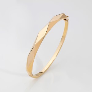May include: A gold bangle bracelet with a geometric design. The bracelet is made of polished metal and has a smooth, shiny finish.