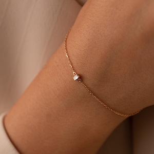 May include: A delicate gold chain bracelet featuring a single, small, rectangular baguette-cut clear gemstone centered on the chain. The bracelet is constructed with fine, polished gold links that form a simple, continuous loop around the wrist.